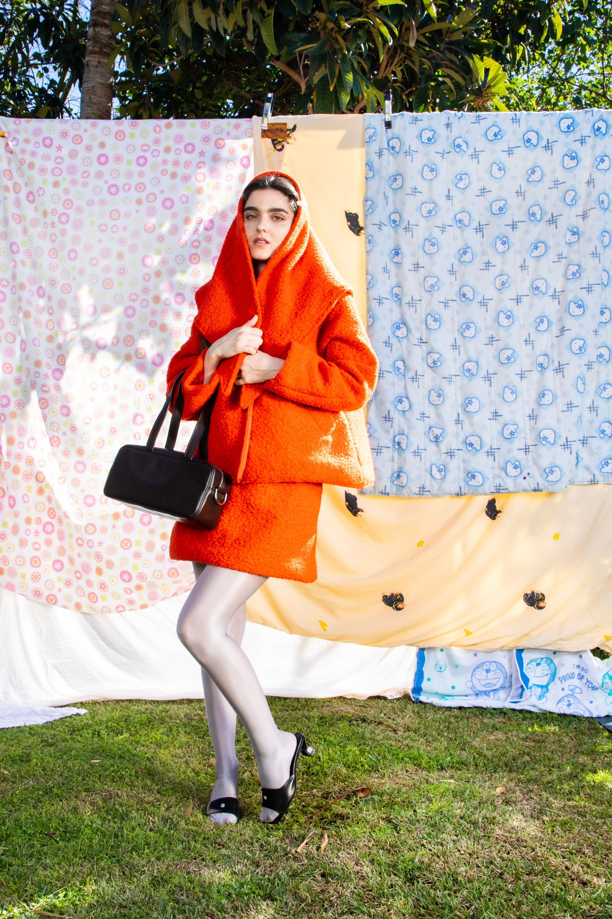 Full body image of a female model in an orange matching coat and scarf set with silver leggings and a black rectangular bag against a backdrop of patterned quilts.